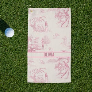 May include: A white golf towel with a pink toile pattern featuring golfers and tropical scenery. The towel has a pink band with the name "OLIVIA" printed on it. A golf ball sits on the green grass next to the towel.