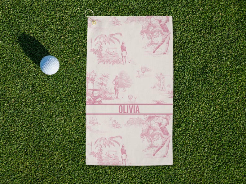 May include: A white golf towel with a pink toile pattern featuring golfers and tropical scenery. The towel has a pink band with the name "OLIVIA" printed on it. A golf ball sits on the green grass next to the towel.