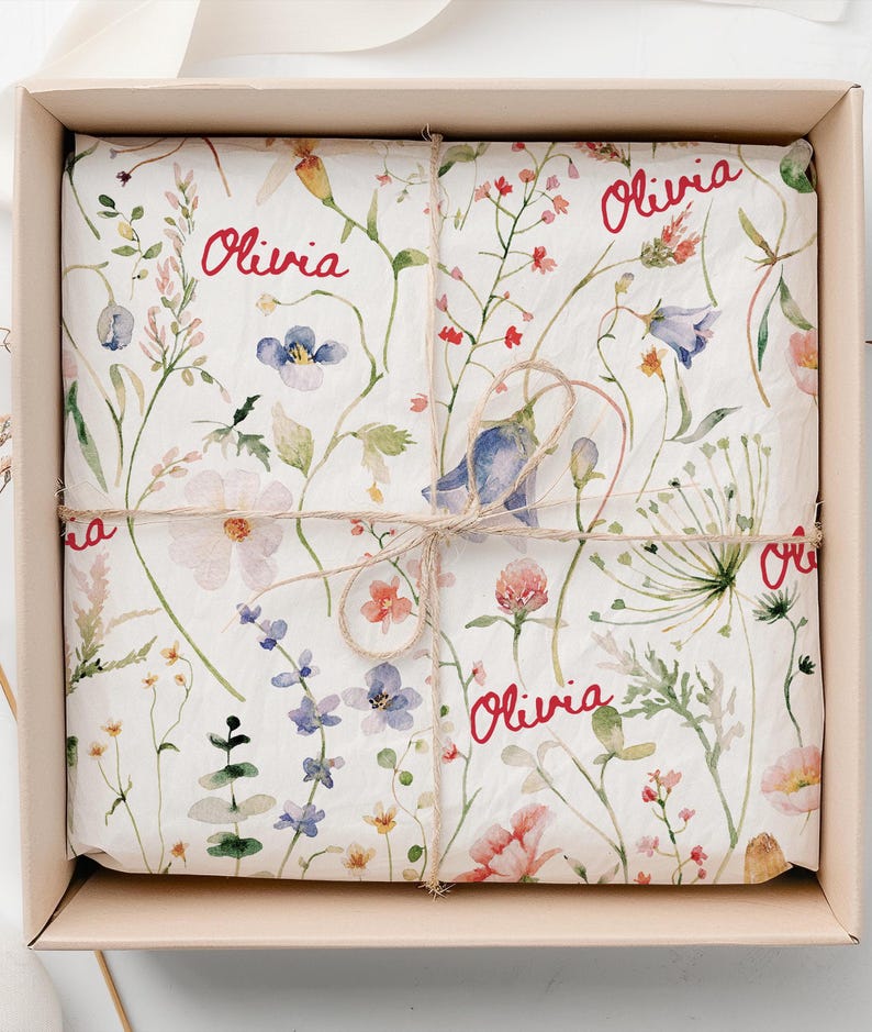 May include: A white gift box with a floral pattern and the name "Olivia" written in red on the wrapping paper. The box is tied with a brown twine.
