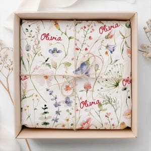 May include: A gift box wrapped in white tissue paper with a floral pattern. The paper has the name "Olivia" written in red cursive on it. The box is tied with twine.