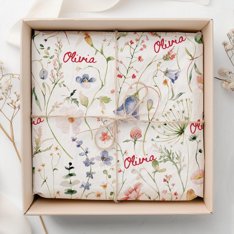 May include: A gift box wrapped in white tissue paper with a floral pattern. The paper has the name "Olivia" written in red cursive on it. The box is tied with twine.