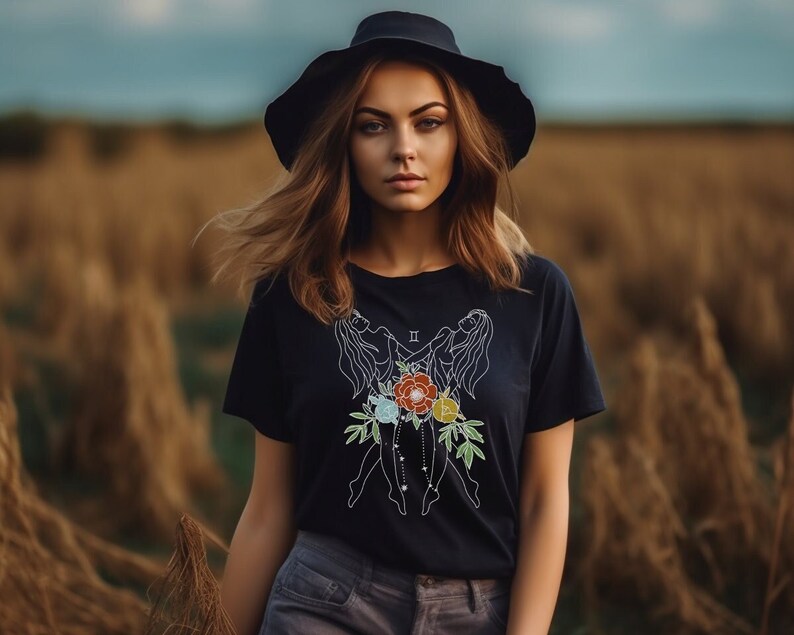 Może przedstawiać: Kobieta w czarnym t-shircie z białym obrysem znaku zodiaku Bliźniąt i kwiatami. Koszulka ma r&oacute;wnież rzymską cyfrę II. Nosi czarny kapelusz i ma długie brązowe włosy.