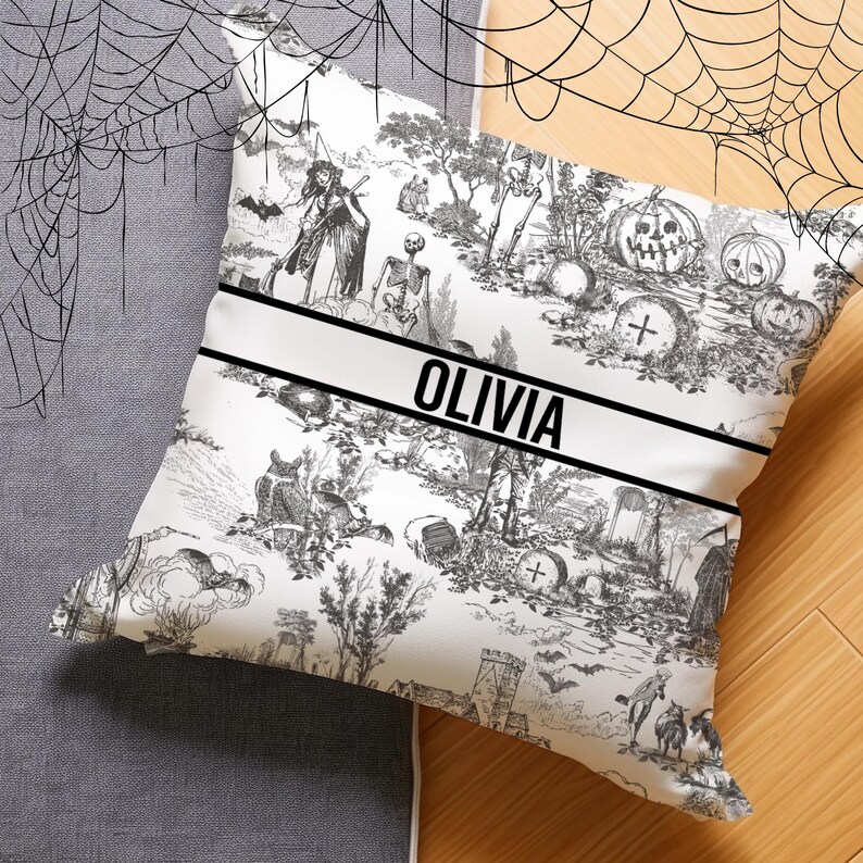May include: Black and white Halloween-themed pillow with a toile pattern featuring a witch, skeletons, pumpkins, and a cross. The pillow has a black and white banner with the name "Olivia" printed on it.