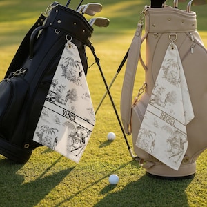 May include: Two golf bags, one black and one beige, stand on a green golf course. Each bag has a golf towel hanging from it. The towels have a black and white toile pattern and the names "VENUS" and "RACHEL" printed on them.