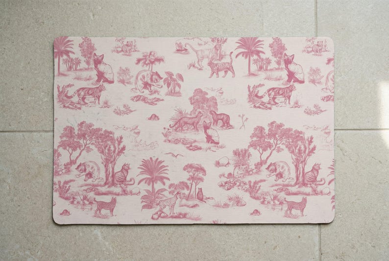 May include: A rectangular placemat in pink and white, decorated with a toile de Jouy design. The pattern depicts various animals, trees, and buildings. The design includes tigers, cats, and other wildlife in a jungle setting.
