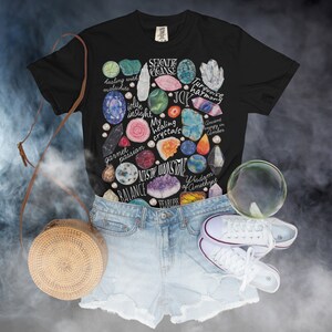 Puede incluir: Camiseta negra con una ilustraci&oacute;n colorida de varios cristales y piedras preciosas. Los cristales est&aacute;n rodeados de texto que describe sus propiedades curativas, como "curaci&oacute;n con malaquita", "intuici&oacute;n de iolite", "armon&iacute;a turquesa", "energ&iacute;a antigua en &aacute;mbar", "sabidur&iacute;a de amatista" y "intr&eacute;pido".