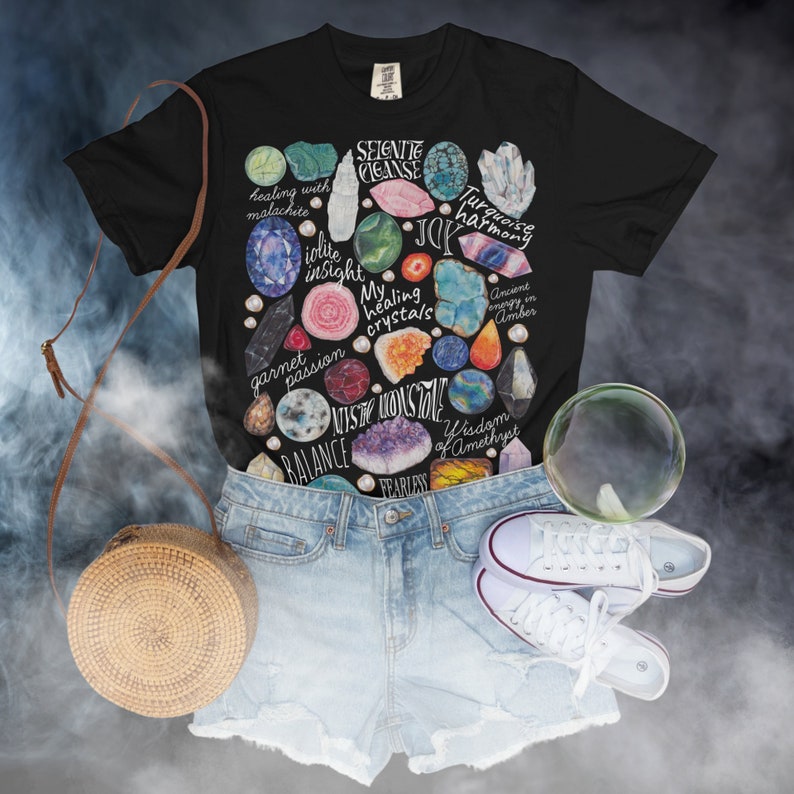 Puede incluir: Camiseta negra con una ilustraci&oacute;n colorida de varios cristales y piedras preciosas. Los cristales est&aacute;n rodeados de texto que describe sus propiedades curativas, como "curaci&oacute;n con malaquita", "intuici&oacute;n de iolite", "armon&iacute;a turquesa", "energ&iacute;a antigua en &aacute;mbar", "sabidur&iacute;a de amatista" y "intr&eacute;pido".