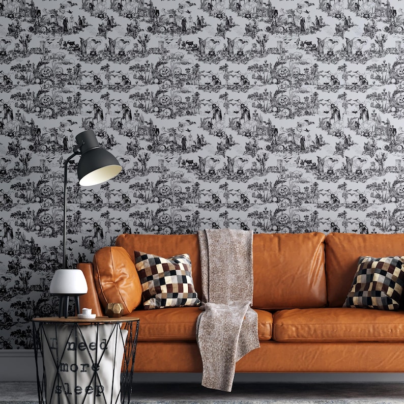 May include: A black and white Halloween-themed wallpaper featuring a toile design with skulls, bats, and figures. The wallpaper is behind a brown leather sofa with decorative pillows and a throw blanket. A black lamp and side table are in the foreground.