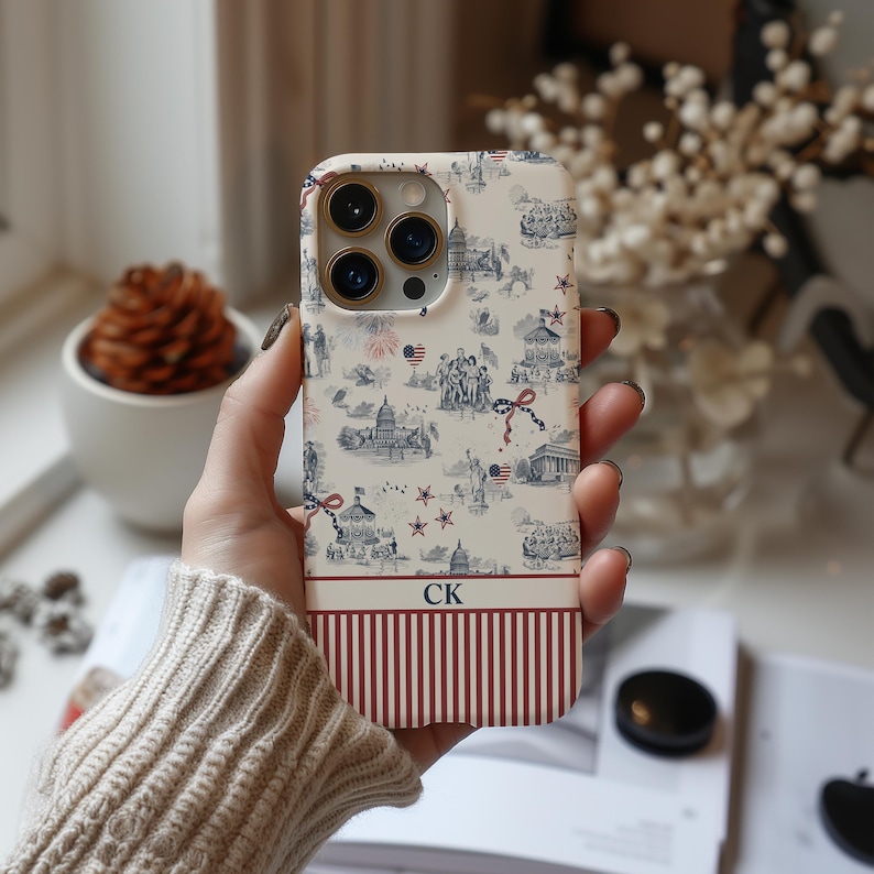 May include: A phone case with a patriotic design. The top portion features a cream background with blue and red illustrations of landmarks, fireworks, and stars. The bottom has red and white vertical stripes. The initials "CK" are at the bottom.