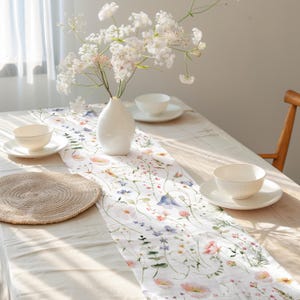 Pu&ograve; includere: Runner da tavolo floreale bianco con un design ad acquerello di fiori di campo, tra cui fiori rosa, blu e viola. Il runner &egrave; su una tovaglia chiara, con ciotole bianche e un vaso di fiori bianchi.