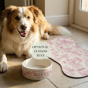 A happy brown and white dog sitting on a tiled floor next to a white bowl labeled "Bella" and a pink dog bed with a floral pattern.