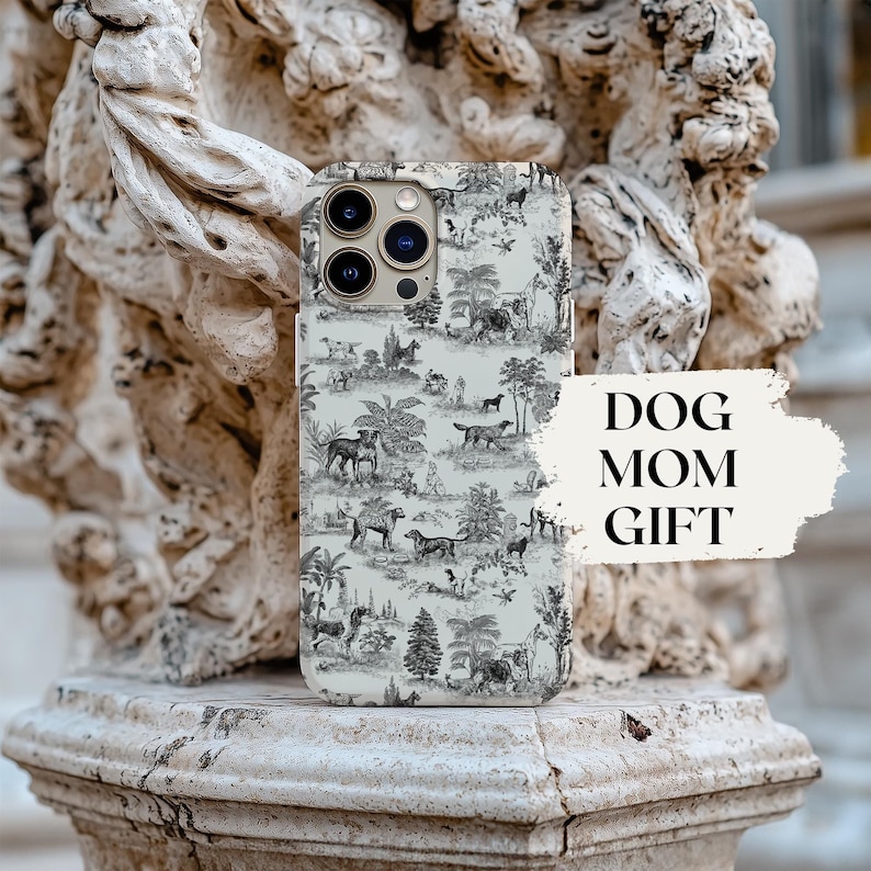 Op de afbeelding: Een witte iPhone-hoes met een zwart-wit toile-patroon met honden. De hoes heeft een heldere camera lens en een zwarte rand. De tekst "DOG MOM GIFT" is zichtbaar op de achtergrond.