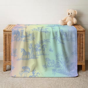 May include: A pastel rainbow-hued blanket featuring a blue unicorn toile design. The blanket is draped over a wicker chest, with a teddy bear on top. The design includes unicorns, trees, and other pastoral elements.