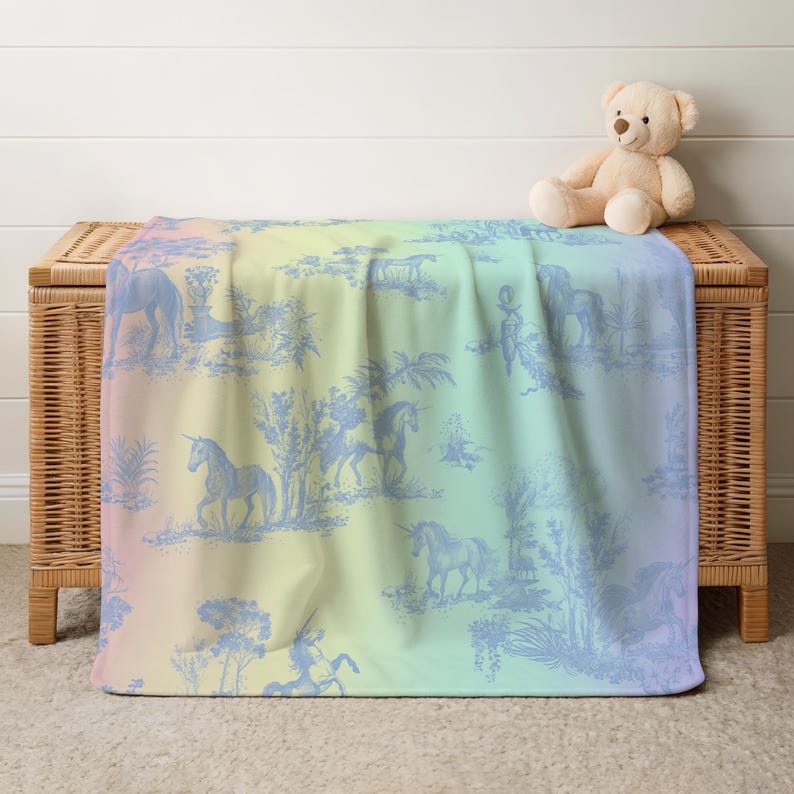 May include: A pastel rainbow-hued blanket featuring a blue unicorn toile design. The blanket is draped over a wicker chest, with a teddy bear on top. The design includes unicorns, trees, and other pastoral elements.