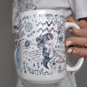 Puede incluir: Taza de cer&aacute;mica blanca con un tema de Acuario, con ilustraciones de una sirena, estrellas y texto manuscrito. La taza tiene un asa grande y est&aacute; decorada con detalles en azul, negro y rosa.