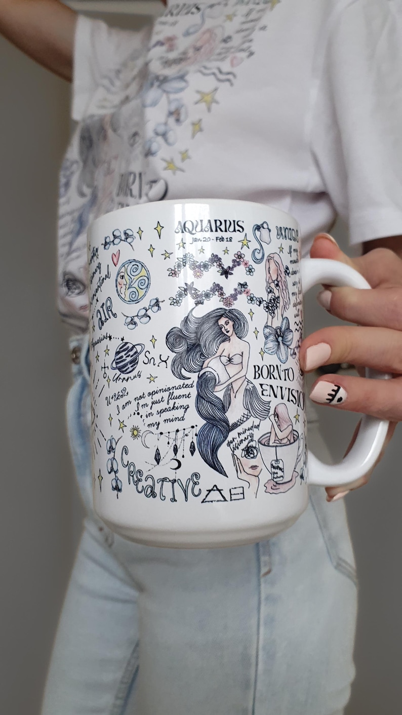 Puede incluir: Taza de cer&aacute;mica blanca con un tema de Acuario, con ilustraciones de una sirena, estrellas y texto manuscrito. La taza tiene un asa grande y est&aacute; decorada con detalles en azul, negro y rosa.