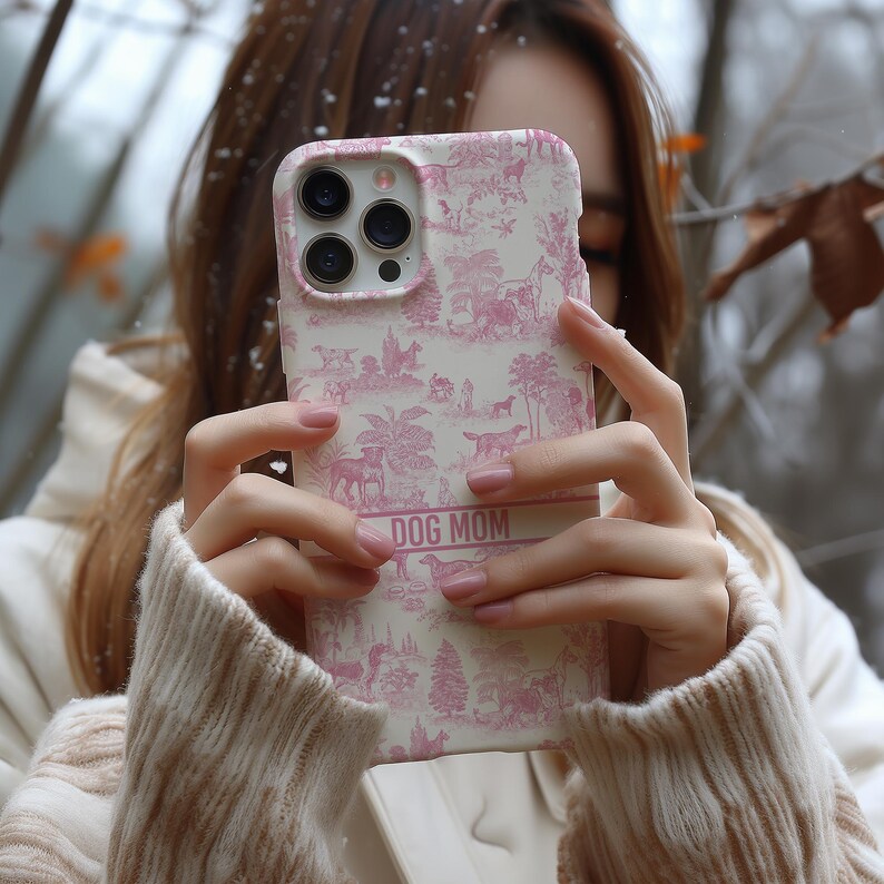 Op de afbeelding: Een witte iPhone-hoes met een roze en wit toile-patroon. De hoes heeft een heldere camera lens en de tekst "DOG MOM" gedrukt aan de onderkant.