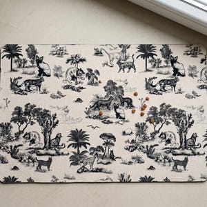 May include: A rectangular beige pet placemat with a black and white toile pattern featuring tigers, trees, and other jungle elements. Several pieces of pet food are scattered on the mat.
