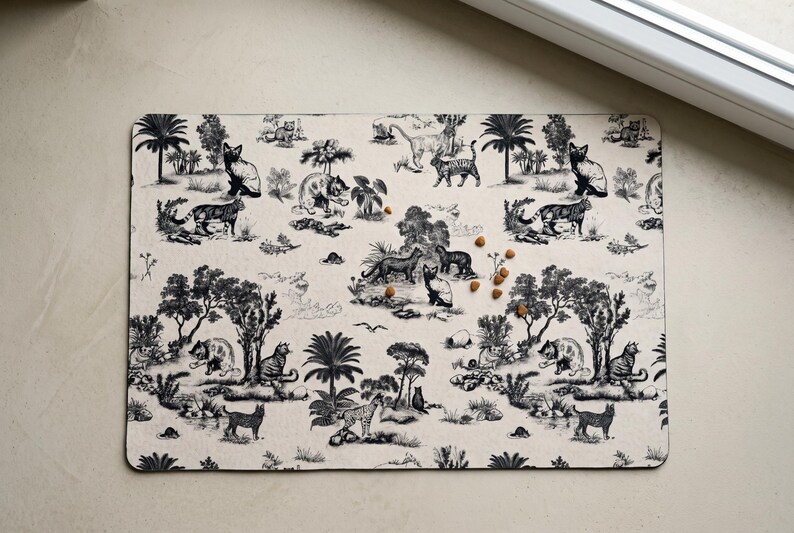May include: A rectangular beige pet placemat with a black and white toile pattern featuring tigers, trees, and other jungle elements. Several pieces of pet food are scattered on the mat.