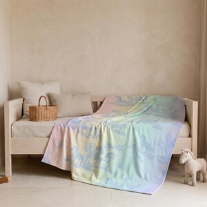 May include: A pastel-coloured blanket with a horse design draped over a light-coloured daybed. The blanket features a gradient of colours, including pink, yellow, and blue. A wooden rocking horse and a stuffed horse are nearby.