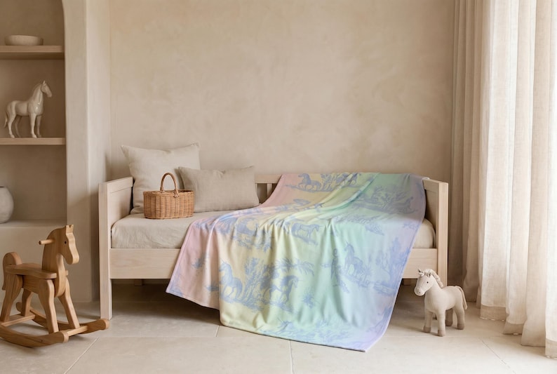 May include: A pastel-coloured blanket with a horse design draped over a light-coloured daybed. The blanket features a gradient of colours, including pink, yellow, and blue. A wooden rocking horse and a stuffed horse are nearby.