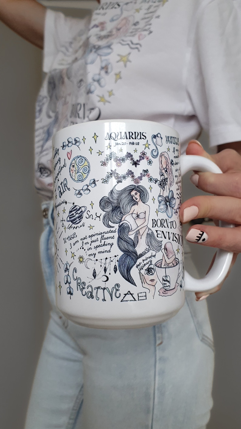 May include: White ceramic mug with a black and white illustration of an Aquarius zodiac sign. The illustration includes a mermaid, stars, and the text "Born to Envision" and "Creative".