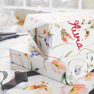 May include: Two gift boxes wrapped in white floral patterned paper. The top box has the name "Olivia" written in red cursive. The boxes are sitting on a white surface with a black ribbon.