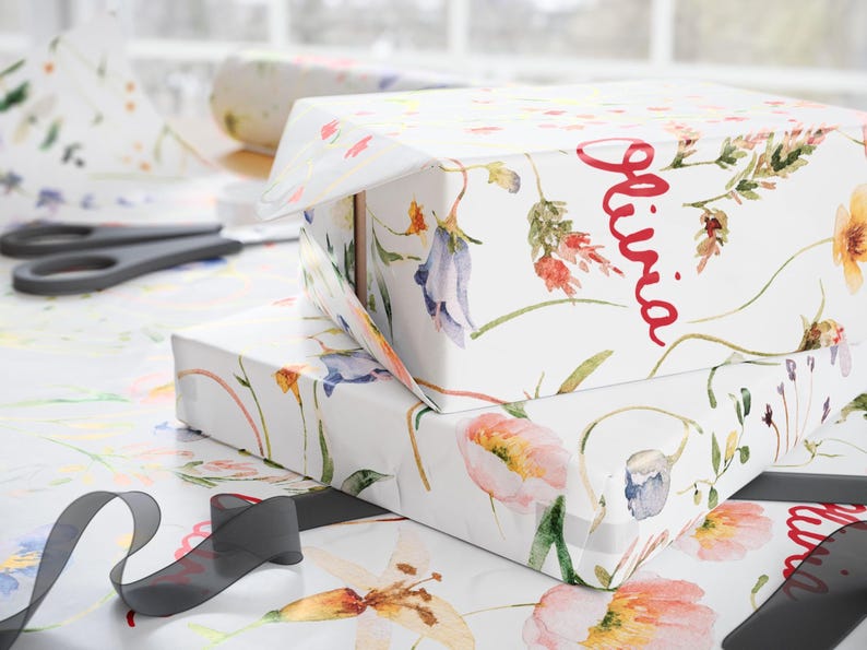May include: Two gift boxes wrapped in white floral patterned paper. The top box has the name "Olivia" written in red cursive. The boxes are sitting on a white surface with a black ribbon.