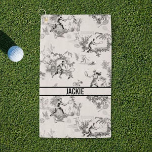 May include: A white tennis towel with a black and white toile pattern featuring tennis players and foliage. The name "JACKIE" is printed in black. A golf ball rests on the green grass next to the towel.