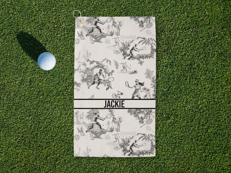 May include: A white tennis towel with a black and white toile pattern featuring tennis players and foliage. The name "JACKIE" is printed in black. A golf ball rests on the green grass next to the towel.