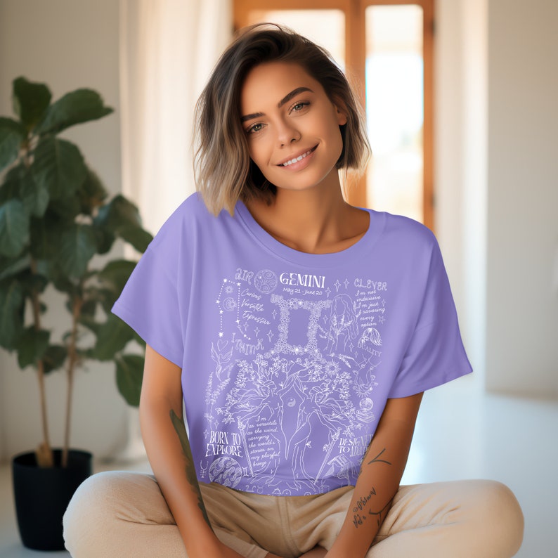 Op de afbeelding: Een lavendelkleurig T-shirt met een wit grafisch ontwerp met het sterrenbeeld Tweelingen, sterrenbeelden en de tekst "AIR GEMINI CLEVER BORN TO EXPLORE DESIRE TO LEARN".