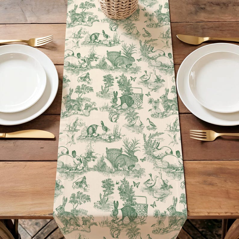a table setting with a green and white table runner with a pattern of rabbits and other animals. There are two white plates and two gold forks arranged on the table.