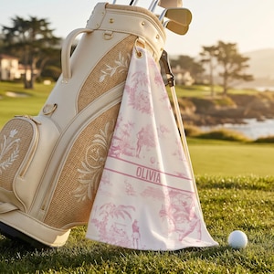 May include: A beige golf bag with a woven texture and embroidered floral designs, paired with a matching towel featuring a pink toile pattern and the name "OLIVIA". A golf ball rests on the green grass.