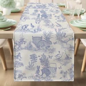 Può includere: Runner da tavolo in toile de Jouy blu e bianco con una scena pastorale con conigli, cesti e farfalle. Il runner è posto su un tavolo di legno chiaro, con posate su entrambi i lati. Il tema è la primavera e la Pasqua.