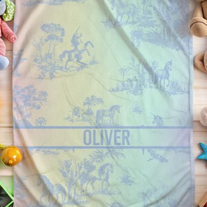 May include: A baby blanket featuring a pastel gradient and a blue unicorn toile design. The name "OLIVER" is printed across the centre. The blanket is surrounded by children's toys, including soft toys and a rattle.