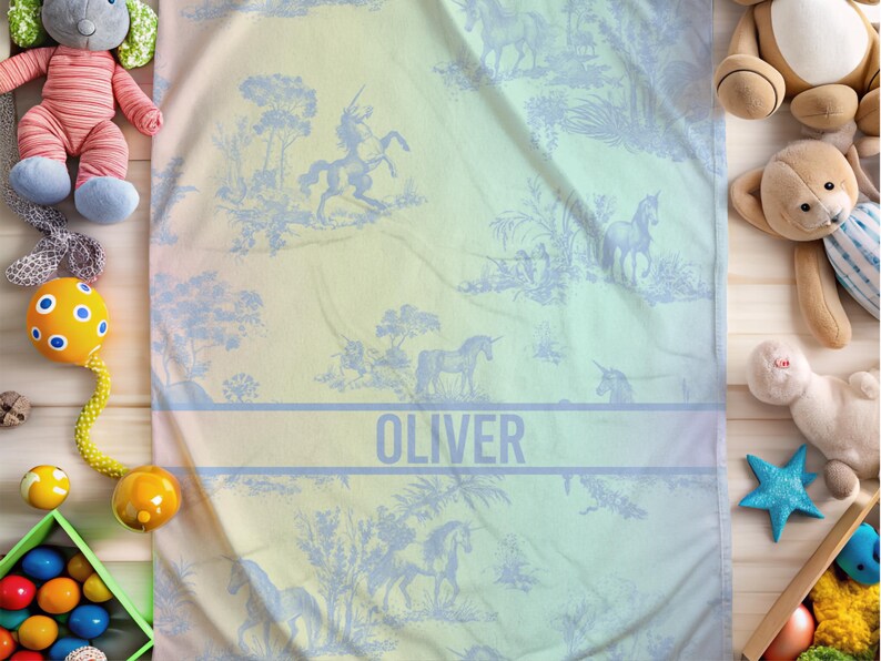 May include: A baby blanket featuring a pastel gradient and a blue unicorn toile design. The name "OLIVER" is printed across the centre. The blanket is surrounded by children's toys, including soft toys and a rattle.