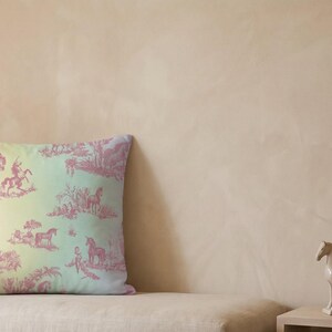 May include: A square cushion with a toile design depicting horses and riders in pink against a gradient background of yellow, green, and blue. The cushion is on a beige sofa.