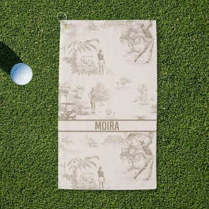 May include: A golf towel with a golf-themed toile pattern in shades of gray and white, with the name "MOIRA" printed across the center. A white golf ball rests on the green grass next to the towel.