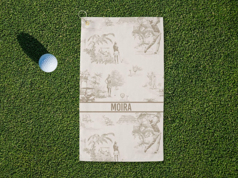 May include: A golf towel with a golf-themed toile pattern in shades of gray and white, with the name "MOIRA" printed across the center. A white golf ball rests on the green grass next to the towel.