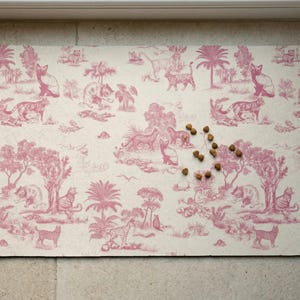 May include: Rectangular pet food mat with a pink toile de Jouy design featuring cats, tigers, and trees on a cream background. Several pieces of pet food are scattered on the mat.