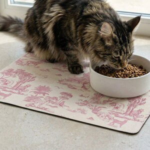 A tabby cat is eating food from a white bowl placed on a pink and white patterned mat.