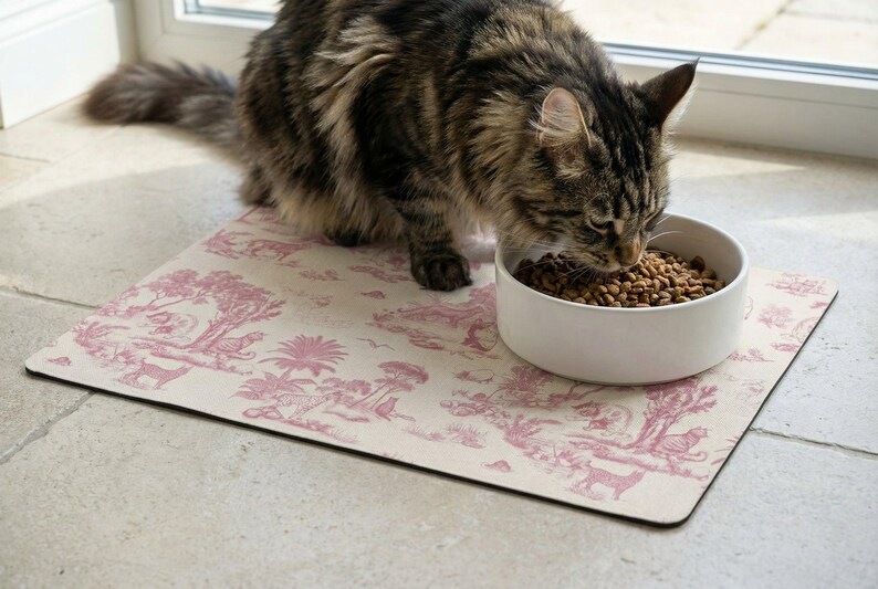 A tabby cat is eating food from a white bowl placed on a pink and white patterned mat.
