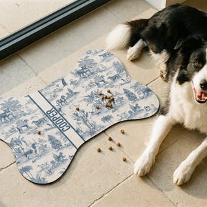 A black and white dog is sitting on a tiled floor next to a bone-shaped dog toy with a pattern of animals and trees. There are several dog treats scattered around the toy.