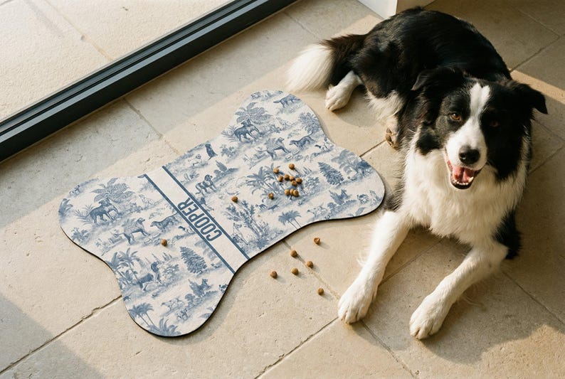 A black and white dog is sitting on a tiled floor next to a bone-shaped dog toy with a pattern of animals and trees. There are several dog treats scattered around the toy.