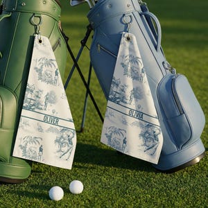 May include: Two golf bags, one green and one blue, stand on a green golf course. Each bag has a matching towel with a blue and white toile pattern and a name printed on it. Two golf balls are on the grass.