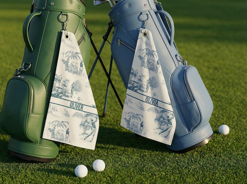 May include: Two golf bags, one green and one blue, stand on a green golf course. Each bag has a matching towel with a blue and white toile pattern and a name printed on it. Two golf balls are on the grass.