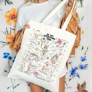 Puede incluir: Una bolsa de tela blanca con un dise&ntilde;o colorido que incluye las palabras "In My Spiritual Era" y varias ilustraciones relacionadas con la atenci&oacute;n plena, el cuidado personal y la espiritualidad. El dise&ntilde;o incluye s&iacute;mbolos como un yin-yang, una flor de loto y un &aacute;rbol con ra&iacute;ces.