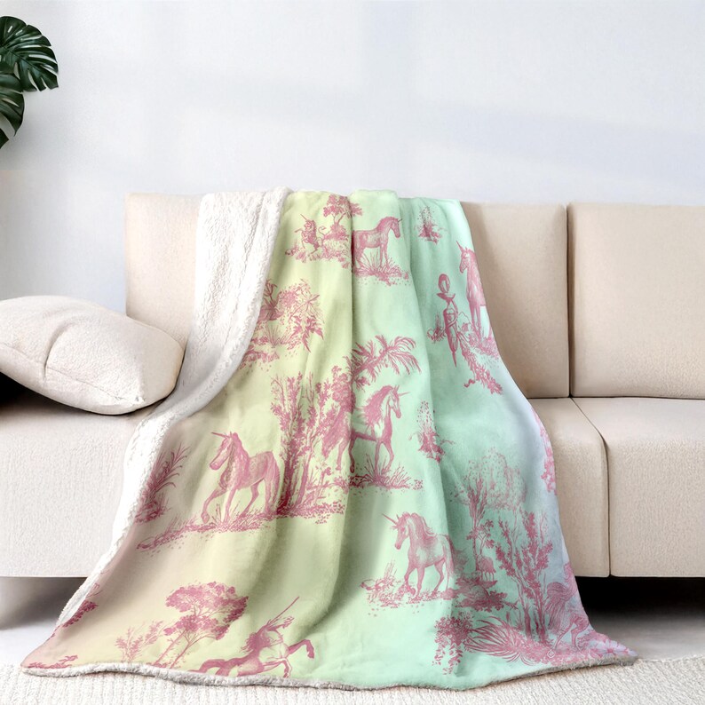 May include: A soft, sherpa-lined blanket with a pink unicorn toile pattern on a yellow and green gradient. The blanket is draped over a beige sofa, displaying detailed illustrations of unicorns and pastoral scenes. The blanket's measurements are in centimetres.
