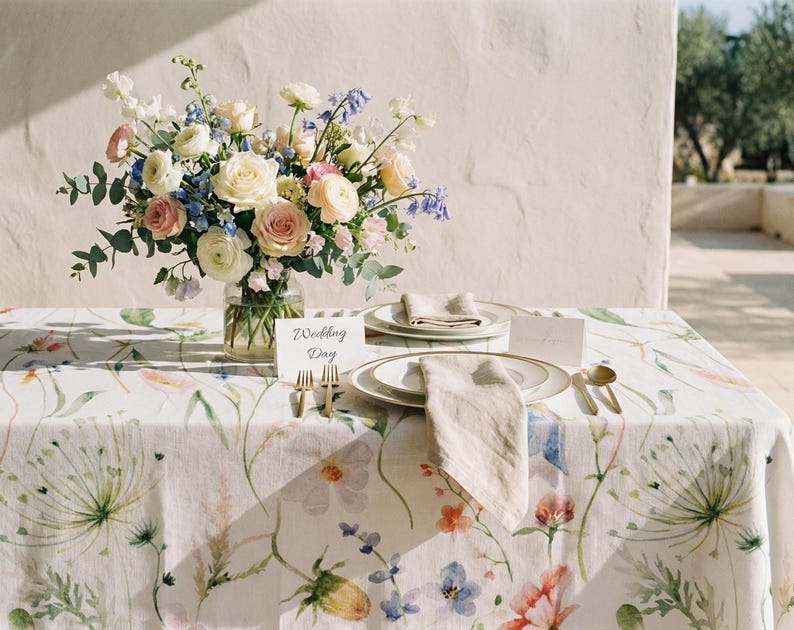 Pu&ograve; includere: Un tavolo nuziale &egrave; apparecchiato con una tovaglia floreale, piatti, tovaglioli e posate dorate. Un vaso di fiori bianchi, rosa e blu &egrave; al centro. Un biglietto con le parole "Wedding Day" &egrave; posto sul tavolo.