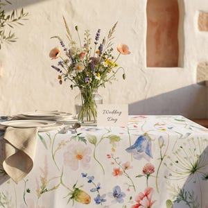 Pu&ograve; includere: Un tavolo apparecchiato per un matrimonio, con una tovaglia floreale bianca, piatti, posate e un tovagliolo piegato. Un vaso di fiori e un biglietto con la scritta "Wedding Day" sono sul tavolo. La scena &egrave; ambientata all'aperto.